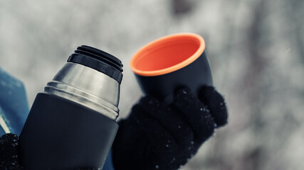 Hiker holds thermos with hot beverage and bright cup in hands wearing gloves at winter forest close up