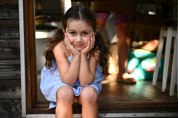 Adorable kid, little girl in blue sundress sits on the doorstep of a country kitchen and smiles...