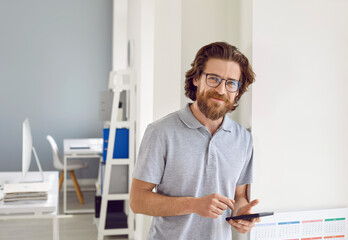 Happy businessman in casual T shirt using mobile phone at work in office. Handsome bearded young man in gray polo shirt standing by window, holding modern smartphone, looking at camera and smiling