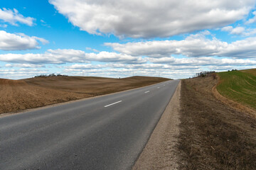 An empty roadway outside the city passes by a rural field and a blooming meadow. Travel by car away from the city and the hustle and bustle. Paved road on a sunny day without cars.