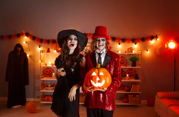 Portrait of happy couple in spooky costumes with Jack-o-lantern. Man in red suit and top hat and...