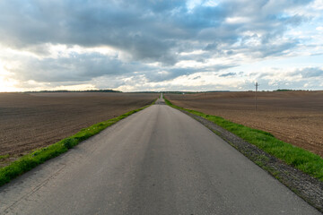 An empty roadway outside the city passes by a rural field and a blooming meadow. Travel by car away from the city and the hustle and bustle. Paved road on a sunny day without cars.