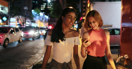 Two female friends walking at night in city, multi-cultural women in sidewalk