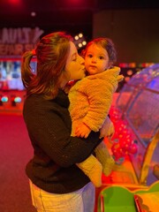 Mom holding a one year old baby in her arms and kissing him in the middle of a colorful playground with countless toys around. Mention the care of parents and fun of children.