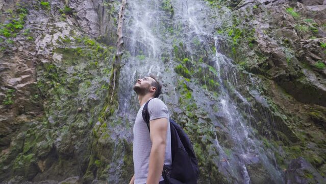 Young man turns around in front of the waterfall.
In front of the waterfall, the young man turns and looks around.
