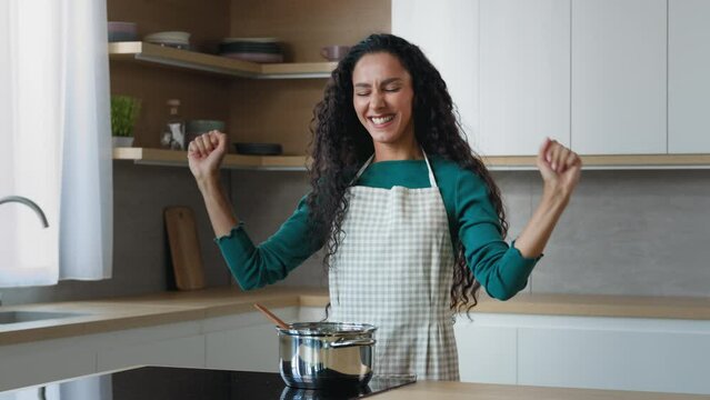 Active funny housewife beautiful woman arabian hispanic maid chef wears apron moving to music rhythm dancing having fun in kitchen preparing dinner cooking for family on stove enjoying culinary time