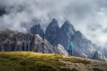 Fototapeta premium Girl with backpack on the hill looking at mountains at sunset in summer. Dramatic landscape with young woman, high rocks, trail, green grass, cloudy sky in Dolomites, Italy. Adventure and hiking