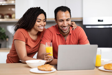 Cheerful young african american family have breakfast, enjoy chat, watching video in laptop in kitchen interior