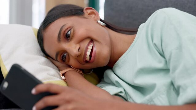 Funny girl relax with social media on phone, smile for communication on app and chat with online friends with smartphone on the sofa in house. Happy, comic and Indian woman reading meme on web