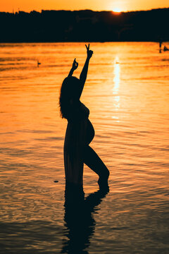 black silhouette of a pregnant woman with a baby bump or belly raising her arms in the air to make a peace or victory sign like a winner or Champion while standing alone in the water of a lake or sea 