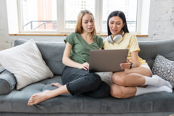 Two millennial diverse girl friends at home watching movies, browsing in the internet, shopping, spending fun time together, chat, talk. Females using technology. Asian and Caucasian model.