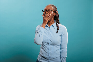 Tired sleepy young adult woman yawning mouth wide open because of fatigue from overtime work. Exhausted african american person catching a yawn while putting hand over mouth. - obrazy, fototapety, plakaty