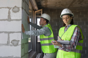 two engineers, a man and a woman in white helmets and protective vests, are standing in the room and holding a plan of the construction site and fire safety in their hands. interior designers