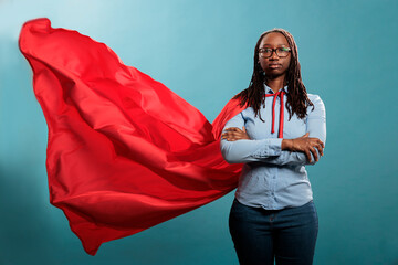 Confident and brave young superhero woman standing with arms crossed while wearing red mighty cape on blue background. Portrait of justice defender posing tough and strong while looking at camera. © DC Studio
