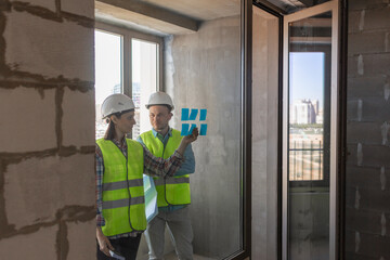 two engineers in protective vests and white helmets glue stickers on the window and discuss the construction plan of the facility or the scheme. European male and female engineers perform fire safety