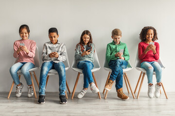 Multiracial Preteen Kids Using Cellphones Sitting Near Gray Wall Indoor