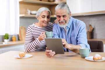 Happy Senior Spouses Using Digital Tablet Together While Having Breakfast In Kitchen