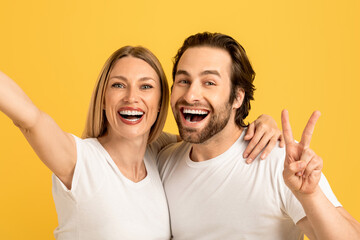 Glad happy millennial caucasian husband and wife in white t-shirts have fun, make peace sign and selfie