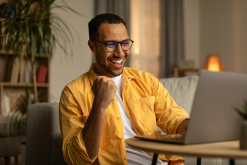 Joyful millennial black man with laptop celebrating online win or work success at home