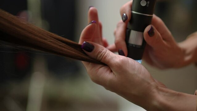 Close-up female hand holding strand moving trichoscope along hair of client in beauty parlor indoors. Unrecognizable Caucasian professional expert trichologist examining hair with equipment