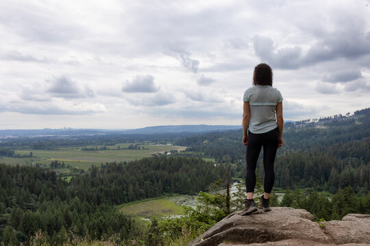 Adventurous Woman Standing on top of a rock overlooking the Canadian Nature Landscape. Minnekhada Regional Park, Coquitlam, Vancouver, British Columbia, Canada.