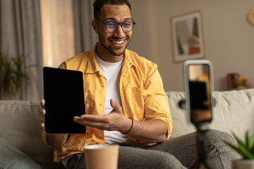 Positive young black male blogger recording video review of new tablet computer on cellphone at...