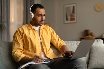 Young black man in headphones having online business conference or webinar on laptop, taking notes...