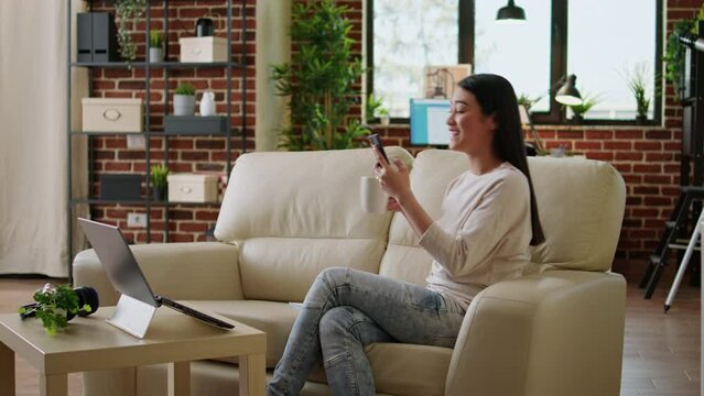 Smiling heartily asian woman watching funny content on smartphone while drinking coffee at home. Joyful young adult person texting with friend while sitting on sofa inside apartment.