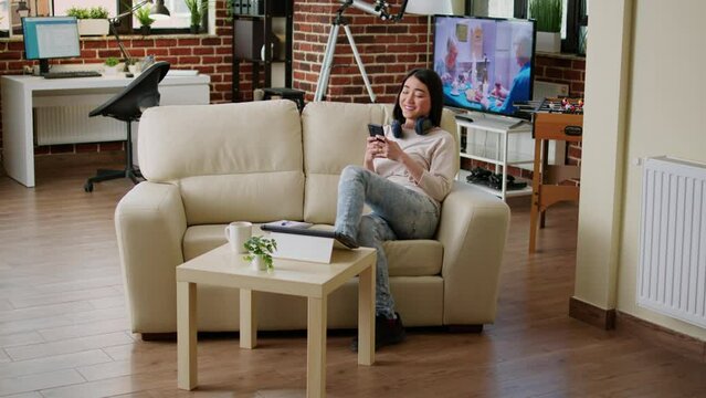 Cheerful woman chatting with friend on modern phone while wearing headphones. Happy young adult person sending messages on smartphone device while sitting on sofa at home.