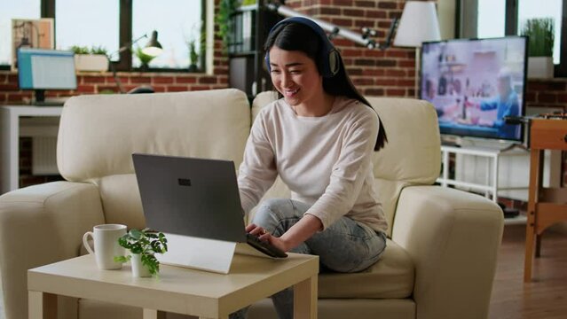 Cheerful woman wearing wireless headphones while working remotely at home on laptop. Happy smiling heartily young adult person doing remote work on modern computer while listening to music.