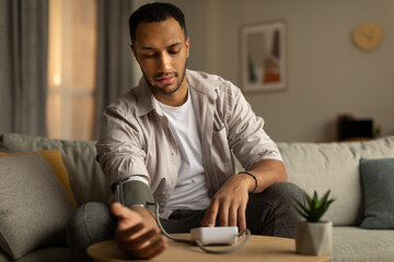 Millennial black man measuring arterial blood pressure, having health problem, sitting on couch at...