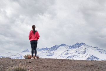 young woman standing in red jacket, looking at snowy mountains in the middle of the Andes Mountains of Chile