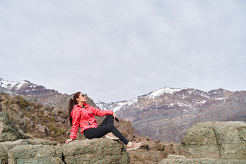 Naklejka na ściany i meble young red haired woman with her hair tied up, wearing a red jacket, sitting on a rock in the middle of the Andes Mountains of Chile