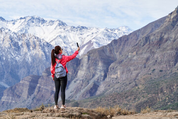 young red haired woman with red jacket and backpack, taking pictures with her phone in the middle of the Andes mountain range in Chile