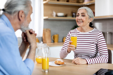 Happy Senior Woman Drinking Juice While Having Breakfast With Husband In Kitchen