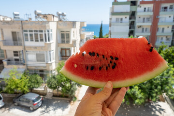 Hand holds piece of watermelon against background of beach town and sea