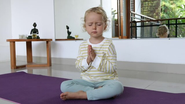 A beautiful blond kid is sitting in meditation on a yoga mat, the child is concentrated on meditation with his arms folded at his chest. Early spiritual development of children in Eastern traditions.