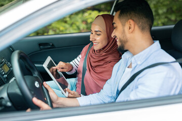 Cheerful millennial muslim female in hijab shows way on tablet with empty screen to husband at steering wheel in car
