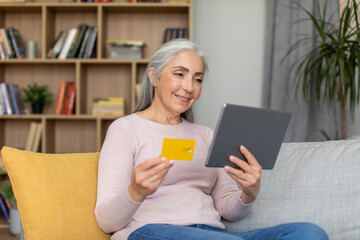 Smiling caucasian old gray-haired female looks at tablet and show credit card in room interior