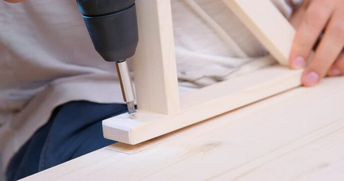 Carpenter screwing a self-tapping screw into a wooden board connects furniture parts. Assembly, manufacturing of handmade wooden furniture. Services by hour. Drill screwing bolt into table, shelf