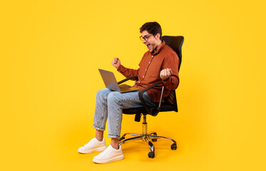 Man Using Laptop Shaking Fists Sitting In Chair, Yellow Background