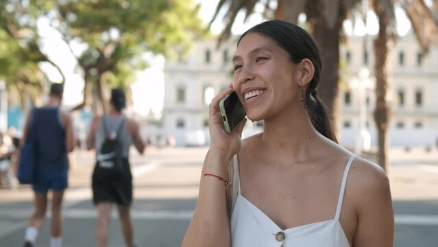 Tracking shot of happy young Latin American woman smiling and talking on cellphone while walking near palms on sunlit street of Barcelona in summer in Spain