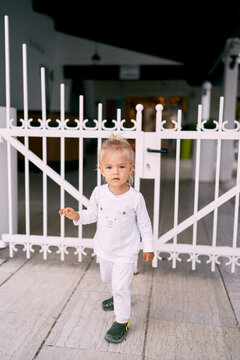 Little girl in a white tracksuit stands in front of a white metal gate on a tile. High quality photo
