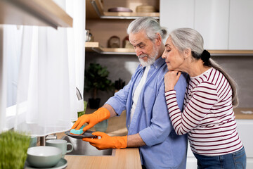 Loving Senior Woman Embracing Her Husband While He Washing Dishes In Kitchen