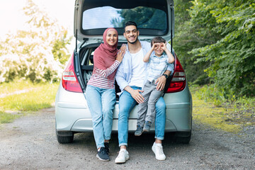 Smiling young middle eastern man and wife in hijab holding baby, sitting in car trunk, resting from trip outdoor