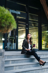 Confident business woman working online on laptop sitting on stairs front of office buildings at downtown. Business persone working remote. Outdoor. Freelancers