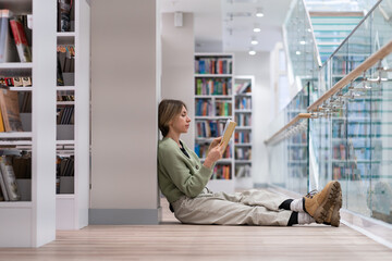 Pensive middle-aged Scandinavian woman book lover spending free leisure time in library, mature female with book in hands sitting in lotus pose on floor near bookcase, enjoying reading. Hobby concept