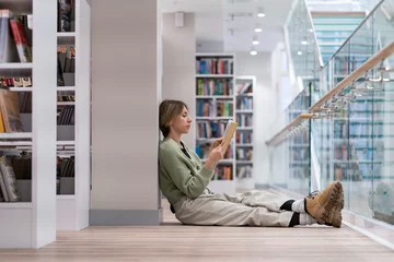 Tuinposter Pensive middle-aged Scandinavian woman book lover spending free leisure time in library, mature female with book in hands sitting in lotus pose on floor near bookcase, enjoying reading. Hobby concept © DimaBerlin