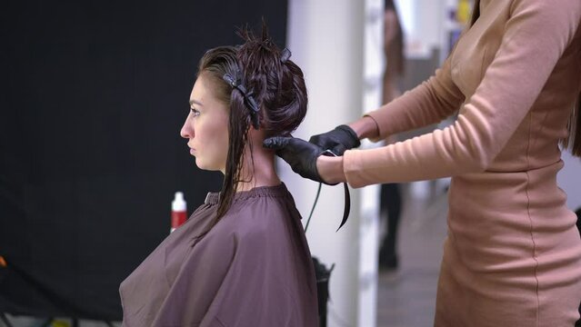 Side view hairstylist using infrared flat iron on wet hair of young client. Unrecognizable professional Caucasian hairdresser straightening strands in slow motion in hair salon