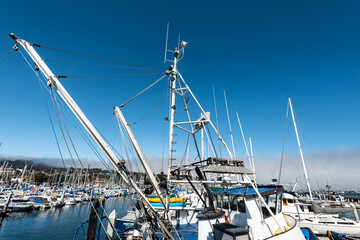 details from masts and equipment of fishing boat docked in marina with many sailboats under blue sky with coastal cloud bank © mariekazalia